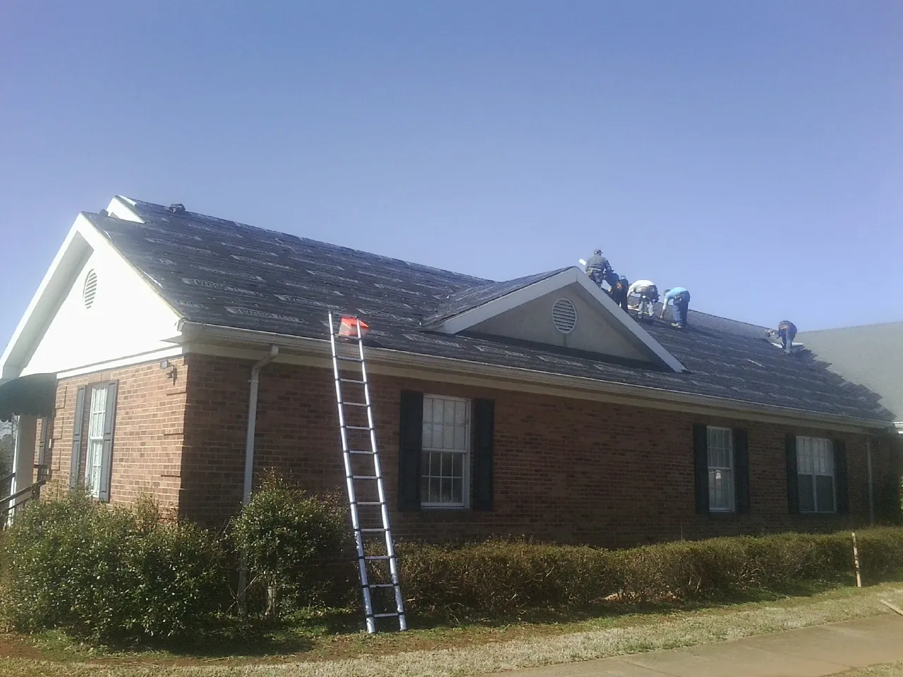 Crew tearing shingles off a brick ranch roof with a ladder set — Metal Roof Repair work by our crew in Locust Grove